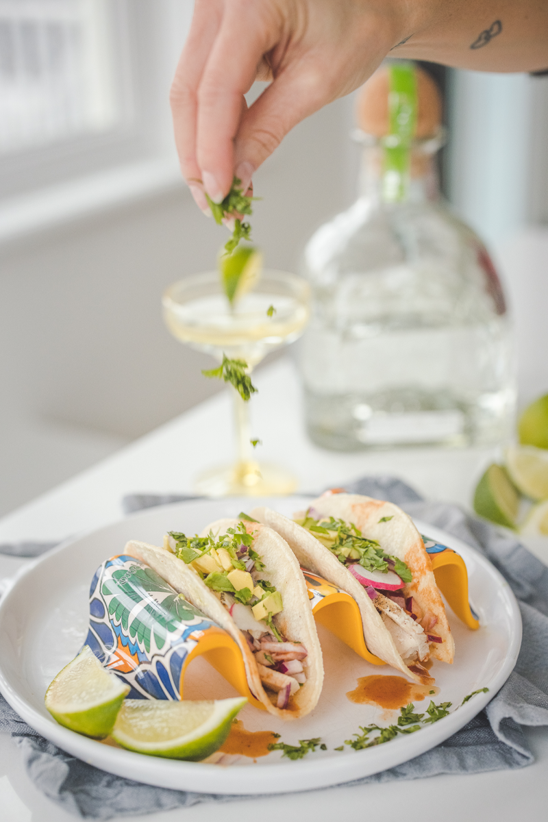 Three tacos on a plate with a bottle of tequila in the background