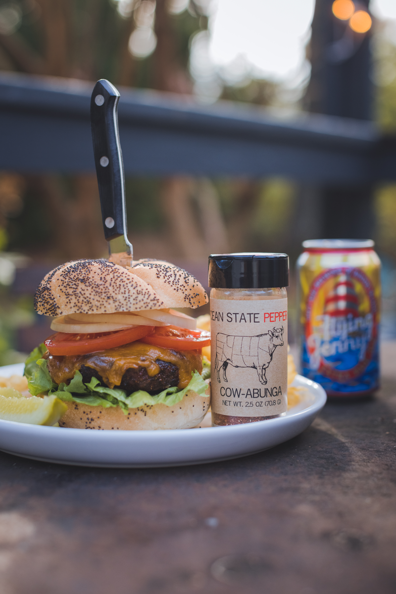 Hamburger on a plate with a bottle of 'Cow-Able' seasoning and a can of beer on a wooden surface.