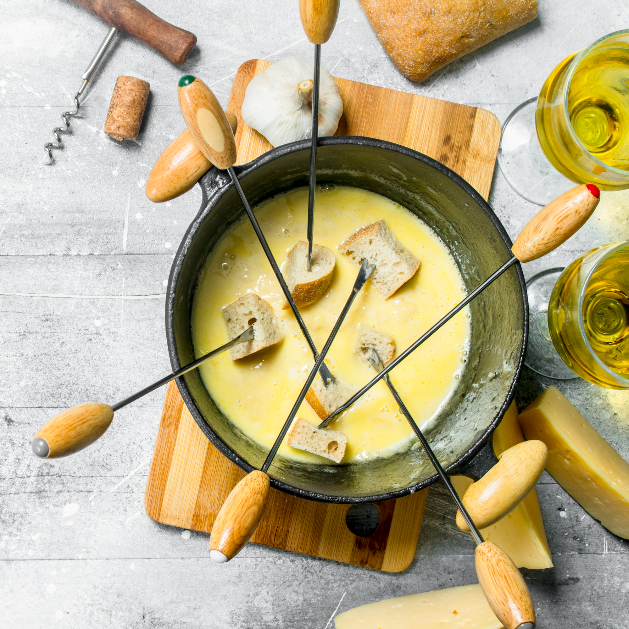 Cheese fondue with bread pieces in a pot on a wooden board, surrounded by wine glasses and bread slices.