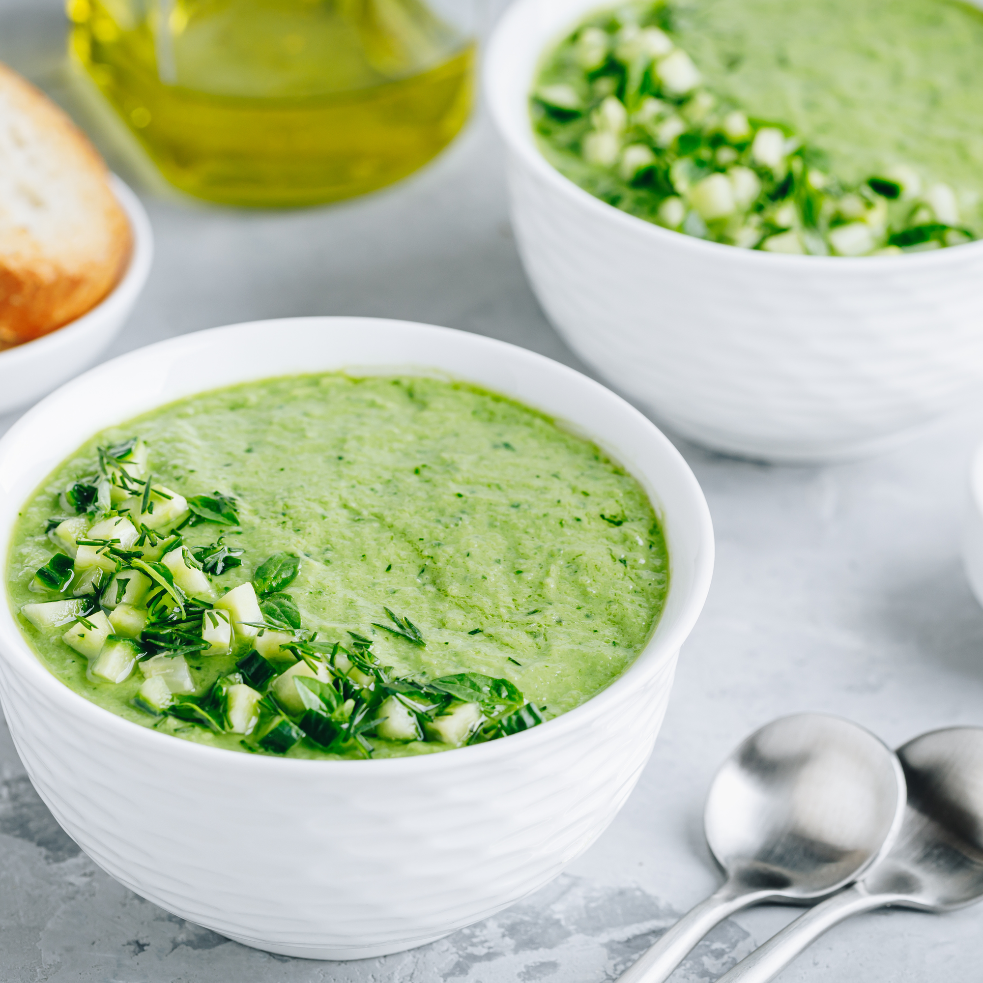 Two bowls of green soup with herbs, a bottle of oil, and bread on a gray surface.